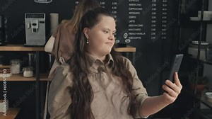 Modern young female barista with Down syndrome working in small cafe recording video for social network on her smartphone