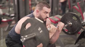 A sportsman sits on bench and lifts a barbell. He pumps lots of iron and weight in order to be fit and build more muscles. He wears a black tank-top, a beard and has a chain with pendant on his neck.