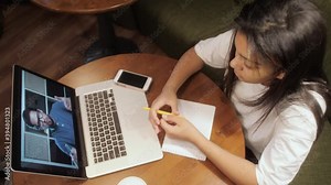 Young girl student watching lesson online and studying from home. Mixed race teenager taking notes while looking at computer screen. Girl student studying at home.