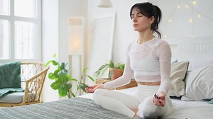 Yoga mindfulness meditation. Young healthy woman practicing yoga in bedroom at home. Woman sitting in lotus pose on bed meditating smiling relaxing indoor. Girl doing breathing practice. Yoga at home