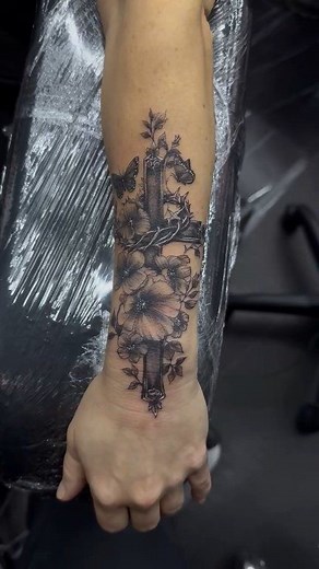Realistic forearm tattoo of a black and grey cross, including a crown of thorns, butterflies, & flowers Performed by Jessie Heart | Outer Beauty Ink