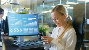 In the Office Attractive Woman Swirles in Chair, Smiles and Uses Mobile Phone. Her Desktop Computer Screen Shows Infographics with Statistical Growth of Her Company's Shares.