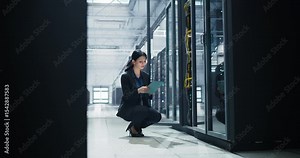 Female Data Center IT Engineer Squatting Down in a Room with Server Racks. Cloud Computing Architect Using Tablet Computer for Servicing the System in Cyber Security and Data Protection Facility