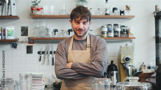 A barista stands at the counter in a cafe with arms crossed. Colleagues move around preparing drinks while a customer waits for their order.