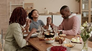 Happy Black family sitting at Easter dinner table, daughter taking hands of mom and dad, they closing eyes and starting praying Stock Video