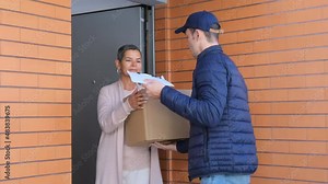 Woman standing in doorway and receiving parcel from courier. Delivery man in uniform and cap delivering cardboard box to customer. Cheerful woman signing for delivery on tablet. Home delivery concept