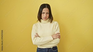 Young, beautiful hispanic woman with crossed arms, standing against an isolated yellow background. her face carries a skeptic, disapproving expression, revealing a negative, nervous persona.