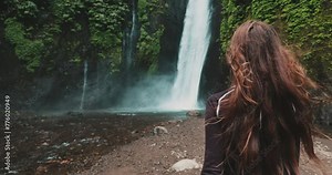 A woman stands at the base of a majestic Bali waterfall, surrounded by lush greenery. Water cascades down the rocks behind her, creating a powerful and mesmerizing scene.