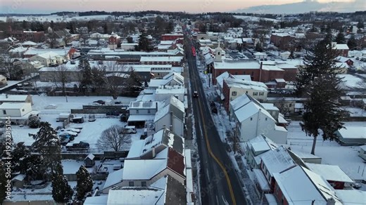 Aerial sunrise view of Manheim, Pennsylvania blanketed in fresh snow, with Main Street lined with historic buildings and quiet residential neighborhoods in peaceful winter setting. approaching.