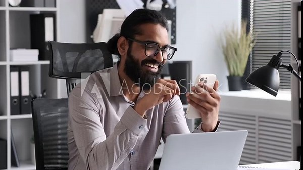 Indian office worker joyfully browses content on phone while at work in the office. outfitted in professional garb, cheerful expressions convey a sense of optimism and enjoyment.