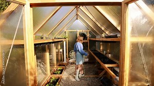 A woman enters her eco-friendly greenhouse, ready to nurture her self-sustaining garden. Living remotely, she embraces a sustainable lifestyle deeply connected to nature