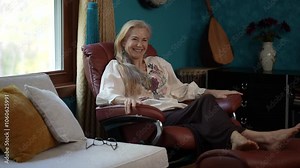 Portrait of mature woman with long blonde hair smiles and relaxes in a brown leather recliner with her feet up. There are flowers in a vase and a musical instrument in the background.