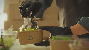 A warehouse worker packs fresh salads into disposable containers in an industrial kitchen. Food delivery employee packing food order into paper container sustainable business idea without waste
