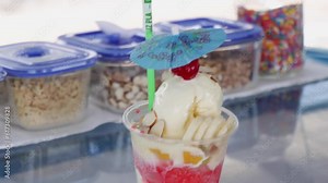 Snow cone with shaved ice, sugar syrup, ice cream, banana fruit and cherry on top, beach dessert served in a plastic cup, with topping containers in the background, Costa Rica