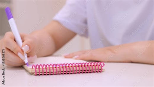 Woman is writing in a pink spiral notebook. She is using a purple marker. The notebook is on a table
