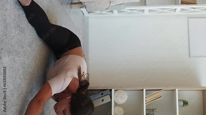 Vertical video. Split exercise. Home stretching. Young flexible fit woman sitting on floor training with spread legs reaching hands forward and to feet in light interior room.