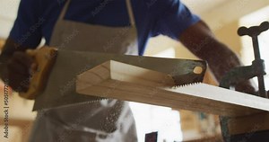 African american male carpenter cutting a wooden plank using a hand saw in a carpentry shop