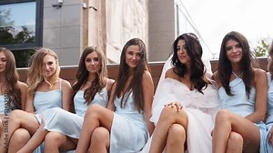 Attractive bride and bridesmaids switching crossing legs sitting on bench seductively in provocative way. Pretty women in identical dresses cross legs simultaneously together flirting at camera.