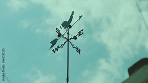 Black metal weather vane on roof of the house. Weathervane cockerel floating under cloudy blue sky . Vintage designed rooster fluger . Arrow rotating by the wind