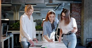 Team of young beautiful women standing over the table and discussing project. IT employees. Successful females talking and brainstorming. Startup concept. Meeting with colleagues.