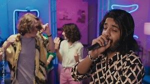 Medium shot of young Middle Eastern guy with beard and long hair singing into microphone at house party, in pink and blue neon light, and male and female friends dancing energetically to rhythm