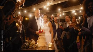 Beautiful Bride and Groom Celebrate Wedding at an Evening Reception Party with Multiethnic Friends. Newlyweds Propose a Toast to Happy Marriage, Standing at a Dinner Table with Cake with Sparklers.