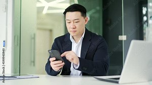 Frustrated asian man having problems with software or service on smartphone while sitting at workplace in modern office. Worried businessman in a formal suit complains about a bad internet connection