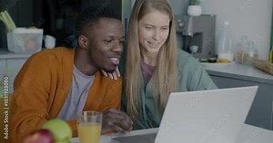 Man and woman young couple using laptop computer browsing discussing online content sitting in kitchen at home. Relationship and modern technology concept.
