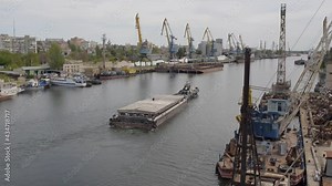 A tug pulls a barge down the river against the backdrop of a port with cranes and moored ships
