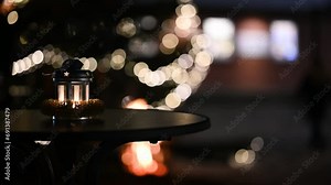 Lantern stands on the table next to a decorated Christmas tree, while a comforting fire flickers in the background. Lantern with festive touch illuminated by festive lights and warm glow of a fire pit