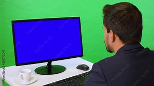 A young businessman sits at a desk and looks at a desktop computer with blue screen - closeup - green screen studio