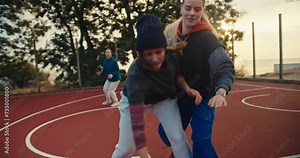 A trio of blonde girls play basketball with an orange basketball on the red basketball court and one of them throws the ball into the basketball hoop during their morning workout