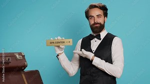 Hotel porter presents direction of spa center using pointer, showing tourists to the right way with wall symbol. Young adult bellhop posing with indicator for relaxation area direction.