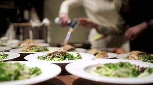 One chef roasts salmon with a gas burner. The second arranges the finished salmon on plates. In the foreground are plates of salad for a group of people. The concept of cooking dinner.