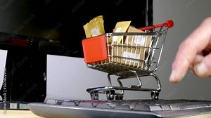 Online shopping concept: Dolly shot of a man’s hand tapping a computer keyboard, with a model shopping trolley full of parcels in the background.