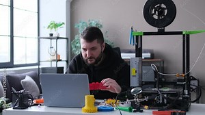 Caucasian man technician working at home workshop with laptop and 3D printer, examining and checking 3D printed model quality. Setting up device. DIY concept.