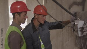 Medium of two male Caucasian builders wearing orange hard hats, standing in building under construction, having discussion, checking electrical panel and sockets