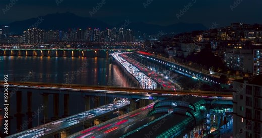 Timelapse displays continuous night traffic light trails streaming across Gangbyeonbuk-ro expressway and Donho Bridge over Han River surrounded by Seoul skyline high-rises and reflections