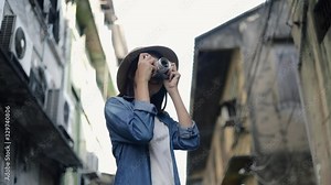 Happy asian female traveler wearing retro fedora hat using film camera taking a photo at a small street in Bangkok Thailand. Enjoying traveling on holiday summer. Solo travel concept.