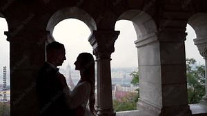 Confident groom meets happy bride for the first time, seeing her in wedding dress, silhouette by the window. Married couple hug and kiss standing near arches and columns of old castle city background
