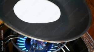 Liquid pancake mix is being poured into a black frying pan. Person swinging the pan around in order to make a flatter pancake.