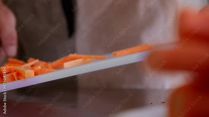 Close-up static shot of cutting carrots. The chef cuts pieces of carrots with a large cleaver and throws them into a plate. The carrot pieces are juicy bright and orange.