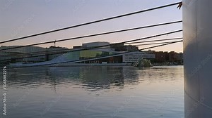 Scenic view of the Oslo Opera House during sunset, with cables crossing the frame and reflecting on calm waters.