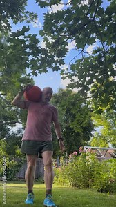 senior, athletic man is exercising in a backyard by walking backwards with a heavy slam ball, green summer scenery