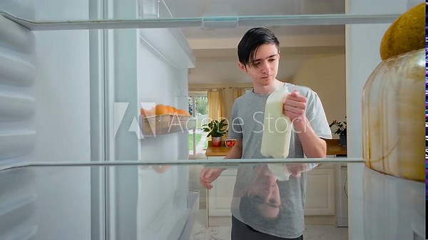 Boy opening fridge and drinking milk straight from bottle, view from inside of fridge. Young thirsty male feeling casual and slightly mischievous. Guy breaking etiquette in private moment, showing