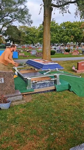 This process is what is referred to as "swinging" a burial vault, also known as "The Wilbert Way" set up. To properly display the vault and to ensure a proper seal, the Burial vault is suspended by cables above the grave. The lid is then separated from the base using lid carriers that roll on lid rails. The weight of the suspended base is what prevents the lid from falling backwards. This particular vault is an Ovation, a display method where the angled lid rails present the lid down toward the 