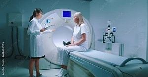 Female doctor is standing near patient and explaining procedure. Female patient dressed up in white is listening to doctor carefully. Medical worker is holding tablet and talk to patient.