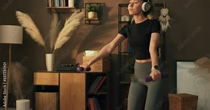 Deep squat. Side view of a young beautiful woman in sportswear doing a squat and holding dumbbells while standing in front of the window listening to music on headphones.
