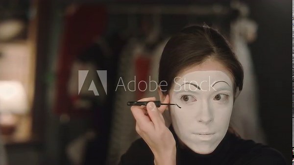 Mime actress drawing black line on white face while applying stage makeup before theatre performance