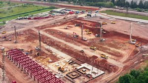 An aerial view of a bustling construction site with heavy machinery in a residential area. Surrounding landscape includes grass, buildings, and asphalt